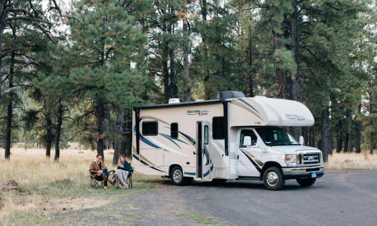 Camping car stationné au milieu des pins, deux personnes à gauche sur des chaises pliantes